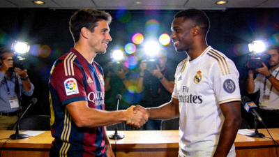 两名身着不同球队队服的足球运动员在新闻发布会上握手，背景是媒体闪光灯