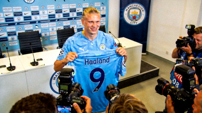 足球明星哈兰德身穿新俱乐部蓝色球衣，在新闻发布会上手持印有自己名字的球衣向媒体展示